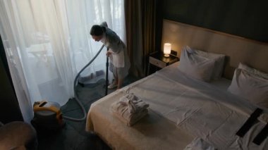 Kadın temizlikçinin rahat otel odasında halı süpürüp temiz bir yatak ve katlanmış havluları...