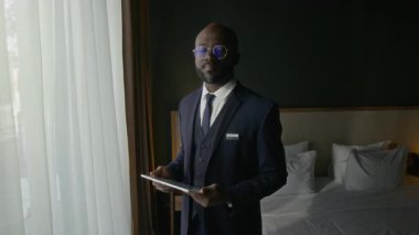 Resmi takım elbiseli Afro-Amerikan otel müdürünün portresi dijital tablet taşıyor ve misafir odasında dururken kameraya gülümseyerek poz veriyor.