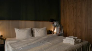 Üniformalı kadın temizlikçi çarşaf ve yastıkları modern otel odasının yakınındaki temiz havlu yığınıyla yeni yapılmış yatağa yerleştiriyor.