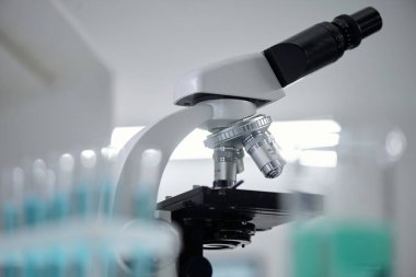 Modern mikroskobun, laboratuarların steril ortamını yansıtan düğmeleri ve lensleri ile yakın çekim görüntüsü. Arka plandaki bulanık test tüpleri aktif bir bilimsel araştırma hissi veriyor