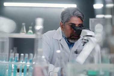 Laboratuvarda yüksek güçlü mikroskop kullanarak örnekleri analiz eden beyaz önlüklü bir bilim adamı. Laboratuvar ekipmanları ve kimyasallarla dolu arka plan bilimsel ortam yaratıyor.