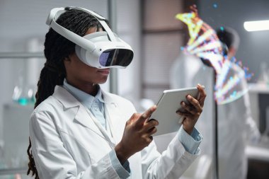 Kadın bilim adamı VR kulaklık takıyor ve laboratuvarda araştırma yapmak için tablet kullanıyor. Arka planda DNA modeli görselleştirme, teknolojiden anlayan çalışma ortamı yaratmak