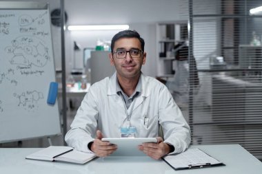 Laboratuvar ortamında, laboratuvar önlüğü giyen, tableti tutan ve arka planda beyaz tahtayla kameraya bakan bilim adamının portresi profesyonel ortam ekliyor.