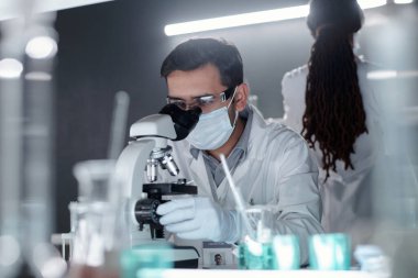 Laboratuvar ortamında bilim adamı, test tüpleri ve laboratuvar ekipmanlarıyla çevrili mikroskopla numuneyi inceliyor.