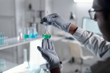 Eldiven giyen bilim adamı modern laboratuvar ortamında bir matarayı tutuyor ve inceliyor. Arka planda çeşitli laboratuvar ekipmanları ve kimyasallar görülebiliyor ve bilimsel bir ortam yaratılıyor.