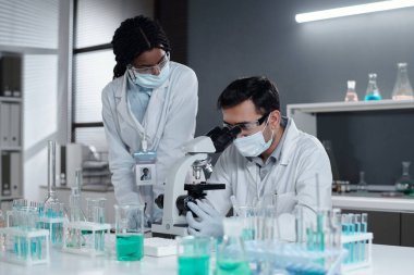 Çağdaş laboratuvar ortamında mikroskop ve renkli test tüpleriyle çalışan bilim adamları laboratuvar önlükleri ve maskeleriyle detaylı araştırma işbirliğine giriştiler.