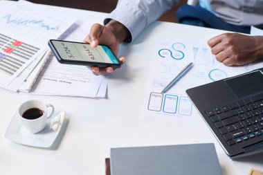 Ofiste oturan beyaz gömlekli, tanınmayan siyah erkek yöneticinin iş günü akıllı telefonunda uygulamayı kullanarak yüksek açılı yakın çekimi.