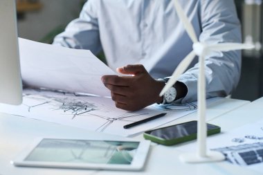 Tanımlanamayan Afrikalı Amerikalı erkek yenilenebilir enerji mühendisi ofisinde oturmuş rüzgar türbini çizimlerine bakıyor.