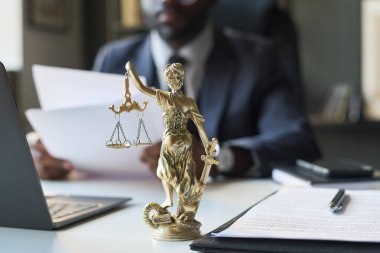 Avukatlık bürosunun masasındaki bronz Adalet Heykeli 'nin arka planında elinde terazi ve kılıç tutan siyah bir erkek avukat ve bulanık arkaplan belgeleriyle çalışıyor.
