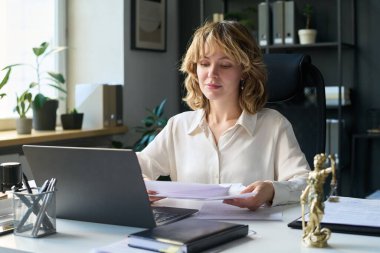 Ofis masasında, üzerinde dizüstü bilgisayar, defter ve bronz heykel olan kağıtlarla çalışan genç bir avukat.