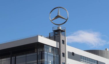 Tanınan marka amblemi olan uzun bir Mercedes-Benz tabelası dışarıda, ağaçların üzerinde duruyor. 