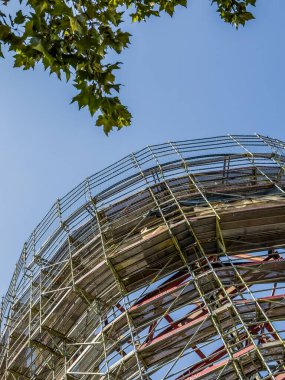 Mavi gökyüzünün altında, köşesinde ağaç dalları olan yuvarlak bir binayı çevreleyen metal iskeleler.