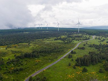 Bulutlu bir günde yeşil enerji üreten rüzgar türbinlerinin hava görüntüsü, Cevennes, Lozere 'in tarım arazisi boyunca bir yol boyunca giden bir araba.