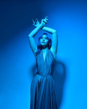 Head and shoulders close up portrait of pretty girl with blue hair wig & elegant gown, with expressive facial expressions & gestural arm poses. colourful neon gel lighting, isolated  studio background