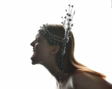 Close up portrait of beautiful girl in side profile, wearing an ornate silver crown headpiece, glowing silhouette lighting on white studio background.