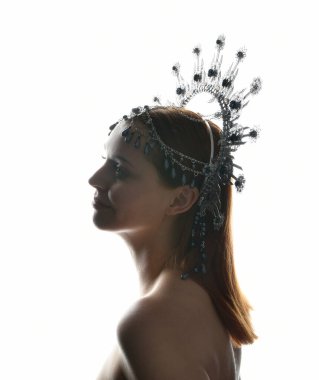 Close up portrait of beautiful girl in side profile, wearing an ornate silver crown headpiece, glowing silhouette lighting on white studio background.