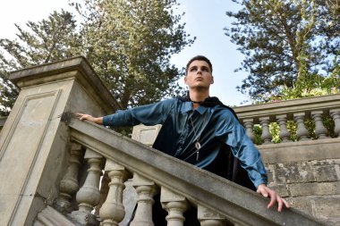 portrait of handsome man wearing fantasy medieval prince costume with golden crown and romantic silk shirt.  sitting on a stone balcony in a historical castle location background.