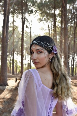Close up portrait of beautiful young blonde model wearing a purple princess fantasy ball gown with flower crown diadem.Pine forest location background with golden lighting.