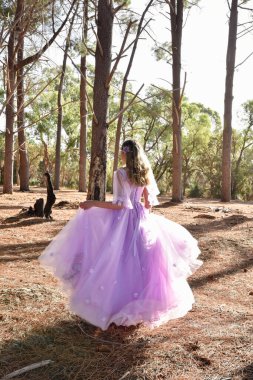 Full length portrait of beautiful young blonde model wearing purple princess fantasy ball gown with flower crown diadem. Walking away posing in pine tree forest location background with golden light