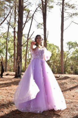 Full length portrait of beautiful young blonde model wearing purple princess fantasy ball gown with flower crown diadem. Walking away posing in pine tree forest location background with golden light