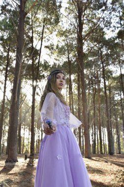 portrait of beautiful young blonde model wearing a purple princess fantasy ball gown with flower crown diadem, posing with magical  crystal wand. pine tree forest location background with golden lighting.