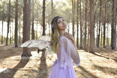 portrait of beautiful young blonde model wearing a purple princess fantasy ball gown with flower crown diadem, posing with butterfly wings. pine tree forest location background with golden lighting.