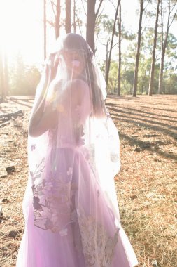 portrait of beautiful young blonde model wearing a purple princess fantasy ball gown with ethereal veil fabric glowing from golden backlit sunshine. pine tree forest location background 