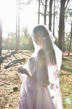 portrait of beautiful young blonde model wearing a purple princess fantasy ball gown with ethereal veil fabric glowing from golden backlit sunshine. pine tree forest location background 