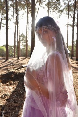 portrait of beautiful young blonde model wearing a purple princess fantasy ball gown with ethereal veil fabric glowing from golden backlit sunshine. pine tree forest location background 