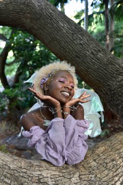 Portrait of beautiful African woman wearing purple fantasy costume, magical fairy wings & flower crown afro, climbing tree branches in forest location with natural glowing lighting.