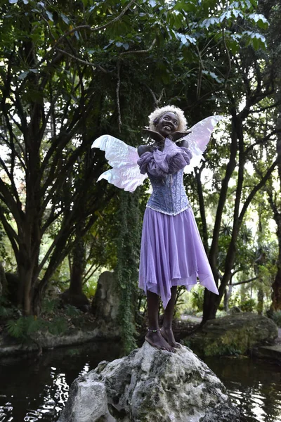 Portrait of beautiful African woman wearing purple fantasy costume, magical fairy wings and flower crown afro, wandering around forest location with natural lighting.