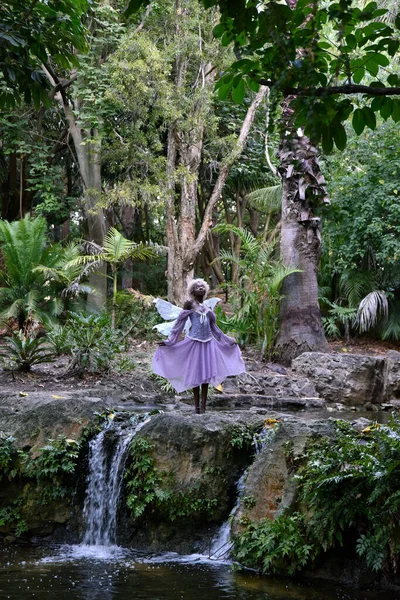 Portrait of beautiful African woman wearing purple fantasy costume, magical fairy wings and flower crown afro, wandering around forest location with natural lighting.