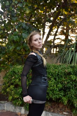 Close up portrait of beautiful female model with blonde plait, wearing black leather catsuit, fantasy assassin warrior.  Posing in forest  tree background