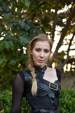 Close up portrait of beautiful female model with blonde plait, wearing black leather catsuit, fantasy assassin warrior.  Posing in forest  tree background