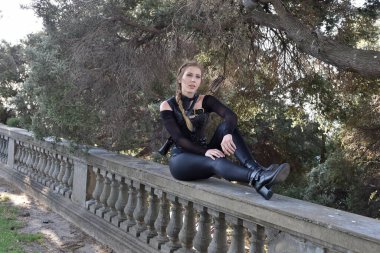 Portrait of beautiful female model with blonde plait, wearing black leather catsuit costume, fantasy assassin warrior.  Crouching sitting pose on stone  balcony of  castle background  clipart