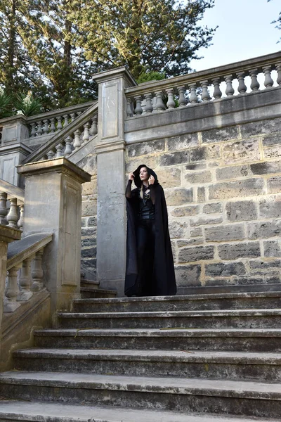 Portrait of beautiful female model with blonde plait, wearing black leather catsuit and flowing hooded cloak, fantasy assassin warrior.  Posing in castle background with stone staircase.