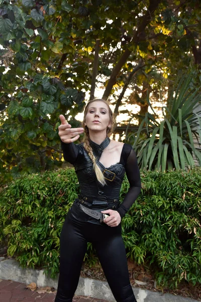 stock image Close up portrait of beautiful female model with blonde plait, wearing black leather catsuit, fantasy assassin warrior.  Posing in forest  tree background