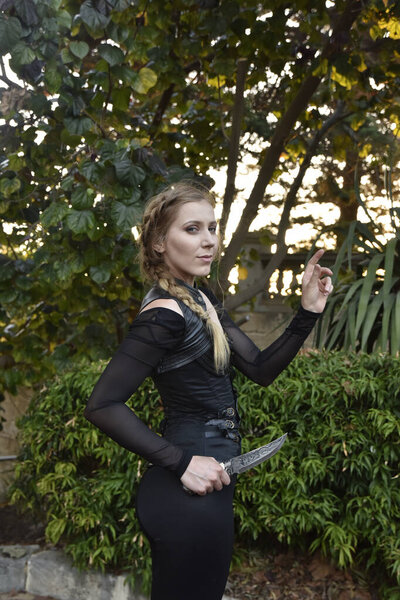 Крупный план портрета красивой женщины-модели с блондинкой косички, в черном кожаном костюме, фантазийный воин-убийца. Поза на фоне леса