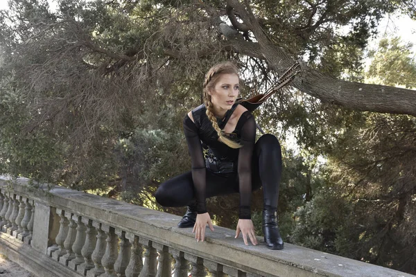 Portrait of beautiful female model with blonde plait, wearing black leather catsuit costume, fantasy assassin warrior.  Crouching sitting pose on stone  balcony of  castle background