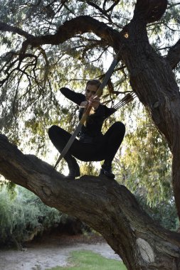 Full length portrait of beautiful female model with blonde plait, wearing black leather catsuit costume, fantasy assassin warrior. holding bow and arrow. Posing in forest  tree branch