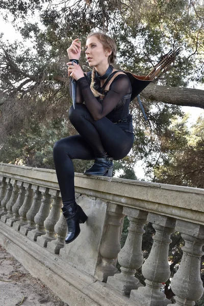 stock image Portrait of beautiful female model with blonde plait, wearing black leather catsuit costume, fantasy assassin warrior.  Crouching sitting pose on stone  balcony of  castle background 