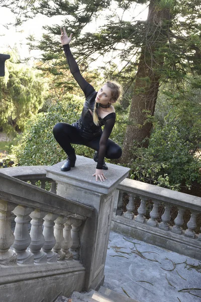 stock image Portrait of beautiful female model with blonde plait, wearing black leather catsuit costume, fantasy assassin warrior.  Crouching sitting pose on stone  balcony of  castle background 