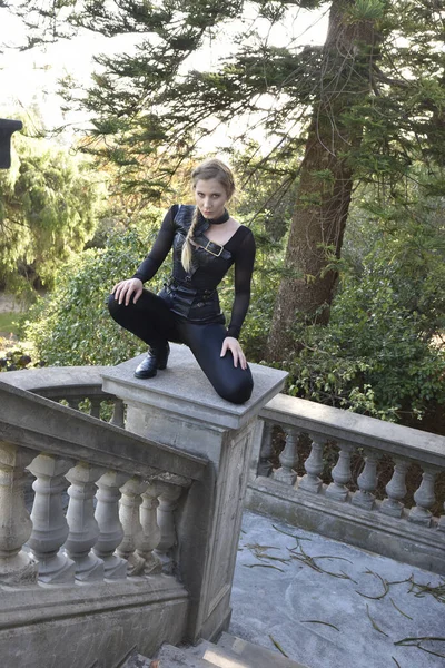 Portrait of beautiful female model with blonde plait, wearing black leather catsuit costume, fantasy assassin warrior.  Crouching sitting pose on stone  balcony of  castle background