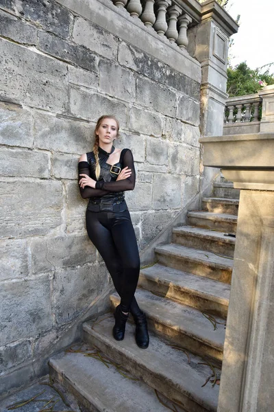 Full length portrait of beautiful female model with blonde plait, wearing black leather catsuit costume, fantasy assassin warrior.  Standing walking pose on stone staircase of  castle background