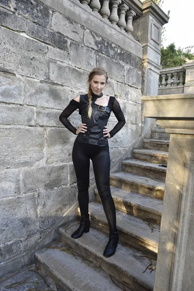 Full length portrait of beautiful female model with blonde plait, wearing black leather catsuit costume, fantasy assassin warrior.  Standing walking pose on stone staircase of  castle background