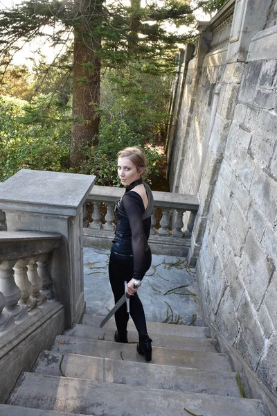 Full length portrait of beautiful female model with blonde plait, wearing black leather catsuit costume, fantasy assassin warrior.  Standing walking pose, holding sword weapon, on stone staircase of  castle background