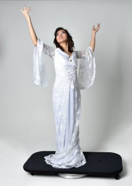 Full length portrait of beautiful woman wearing  fantasy costume, white bridal gown.  standing pose,  gestural arm movements casting a spell. Isolated white studio background. 