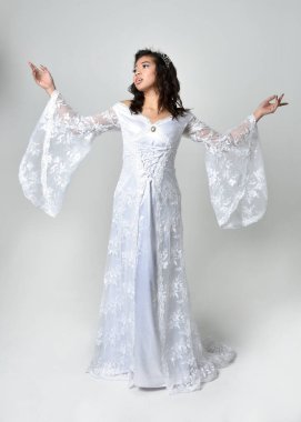 Full length portrait of beautiful woman wearing  fantasy costume, white bridal gown.  standing pose,  gestural arm movements casting a spell. Isolated white studio background. 