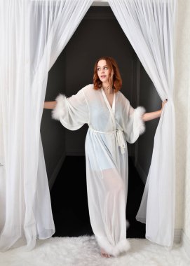 Portrait of beautiful red haired woman wearing glamorous  silk  bridal dressing gown. Standing pose in front of white curtains in bedroom background. 