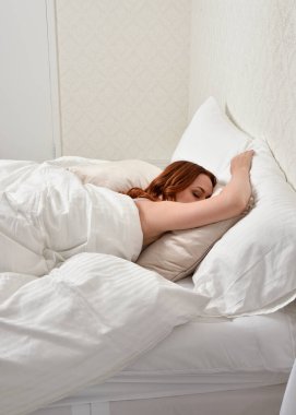Portrait of beautiful red haired woman wearing comfortable pyjamas, casualy resting in bed, getting ready for sleep,  in a glamorous white bedroom location background.